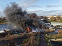 Momento em que um grande incêndio atinge vários veículos pesados e reboques nas docas, enviando nuvens de fumaça para o céu